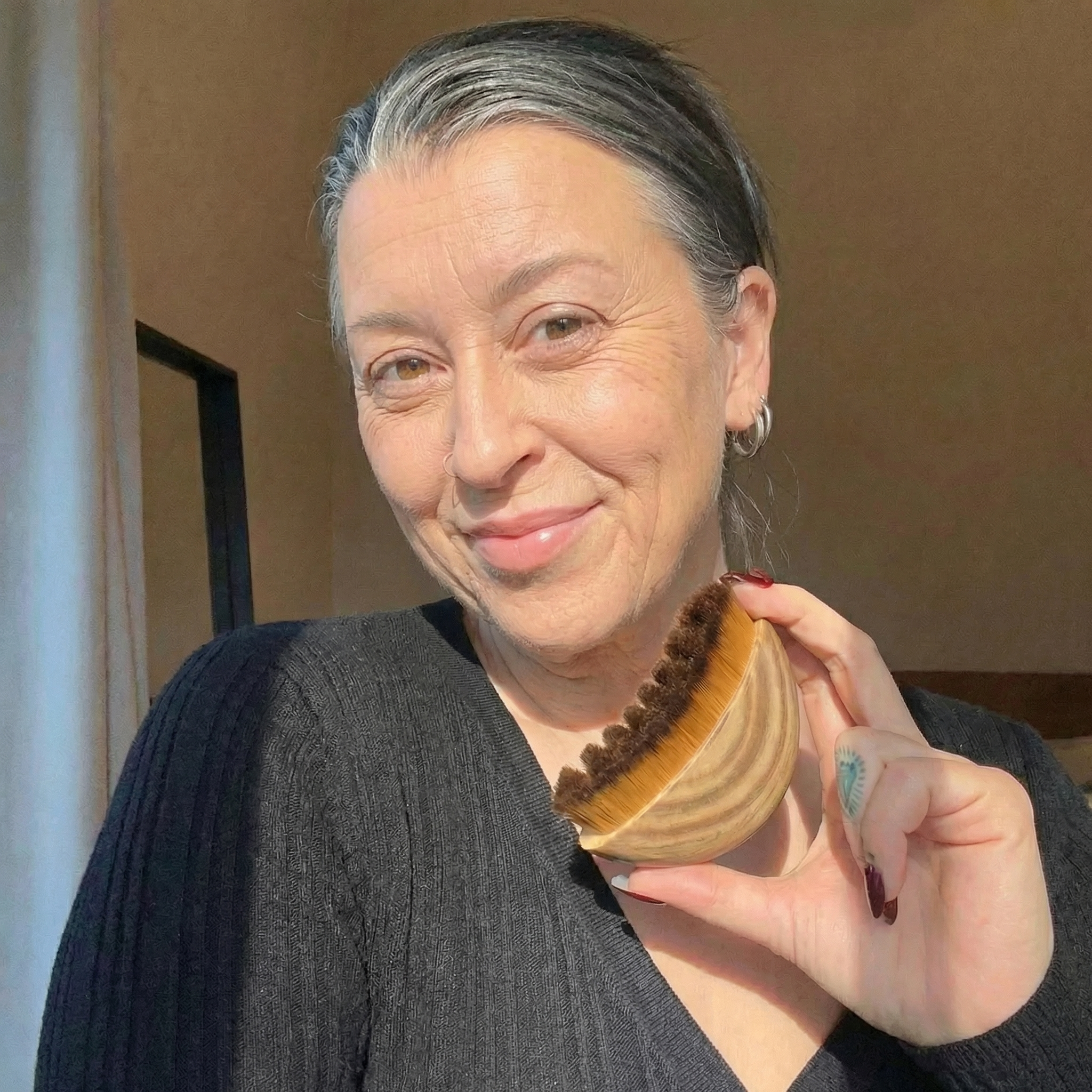 A woman with graying hair pulled back smiles while holding a large, rounded makeup brush.