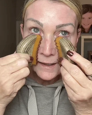 A woman holds two large, ridged, shell-like objects with serrated amber edges on either side of her nose.