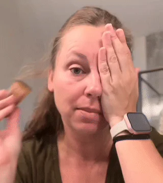 Person applying makeup with a brush, covering one eye with a hand.