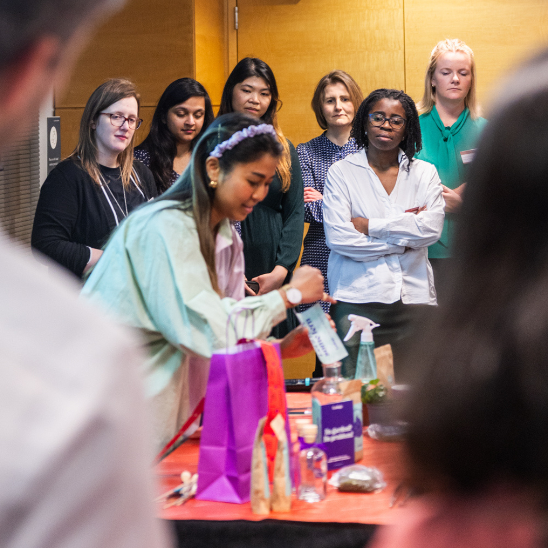 A group of people watching a woman demonstrate something on a table with various items.