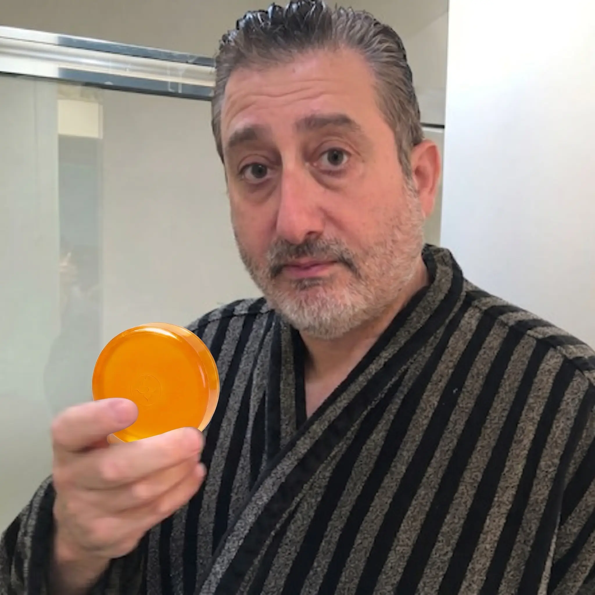 A man in a striped bathrobe holds up a round, translucent orange bar of soap.