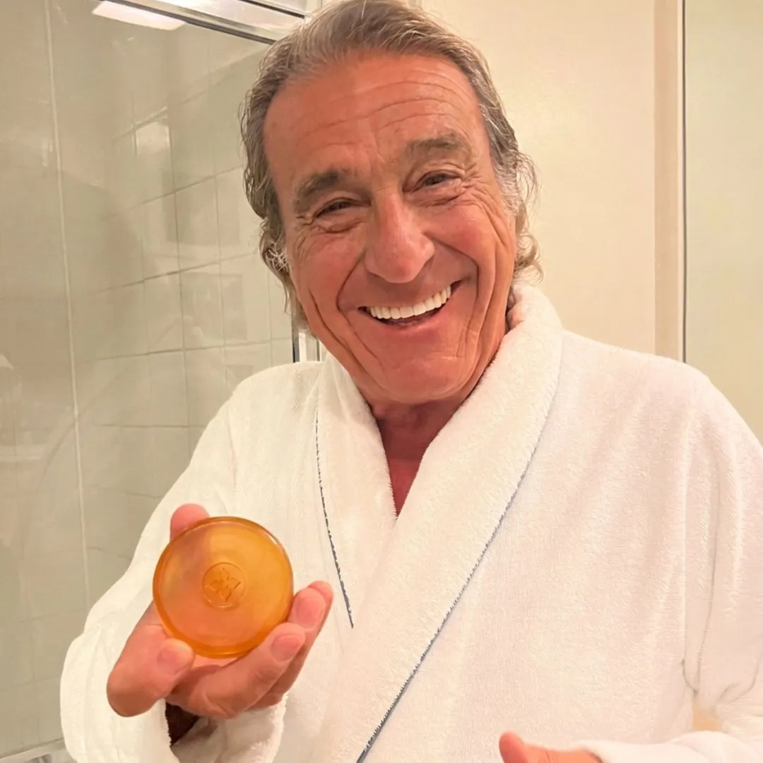 A smiling older man in a white bathrobe holds up a round, amber-colored bar of soap.