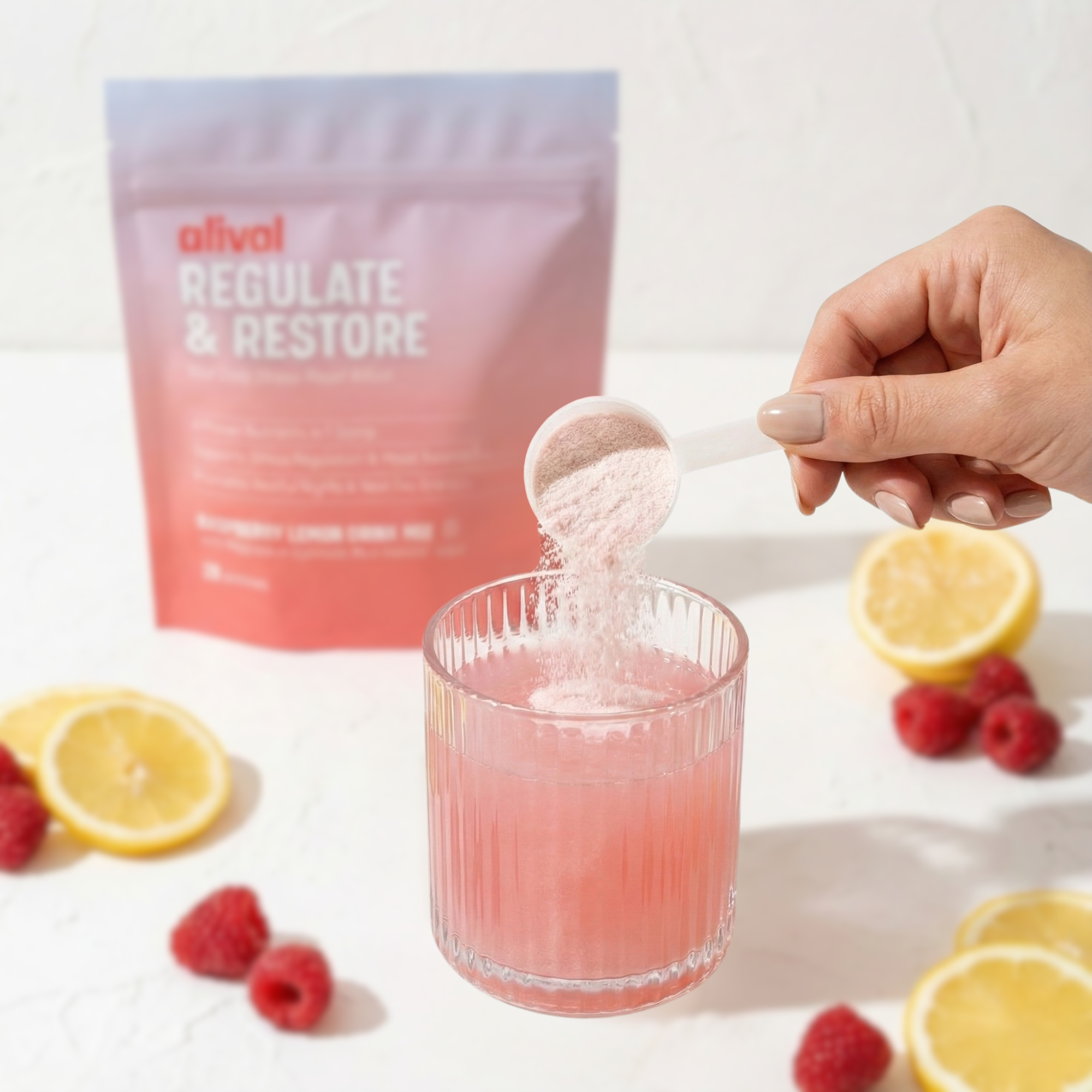 A hand pours pink supplement powder into a glass, with the product bag, raspberries, and lemons nearby.