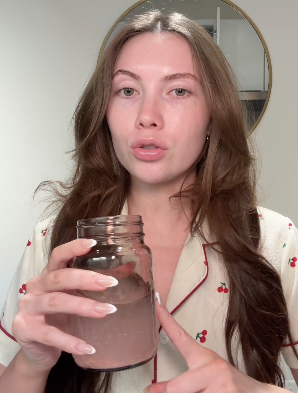 A woman in cherry-print pajamas holds a glass jar with a cloudy liquid inside.