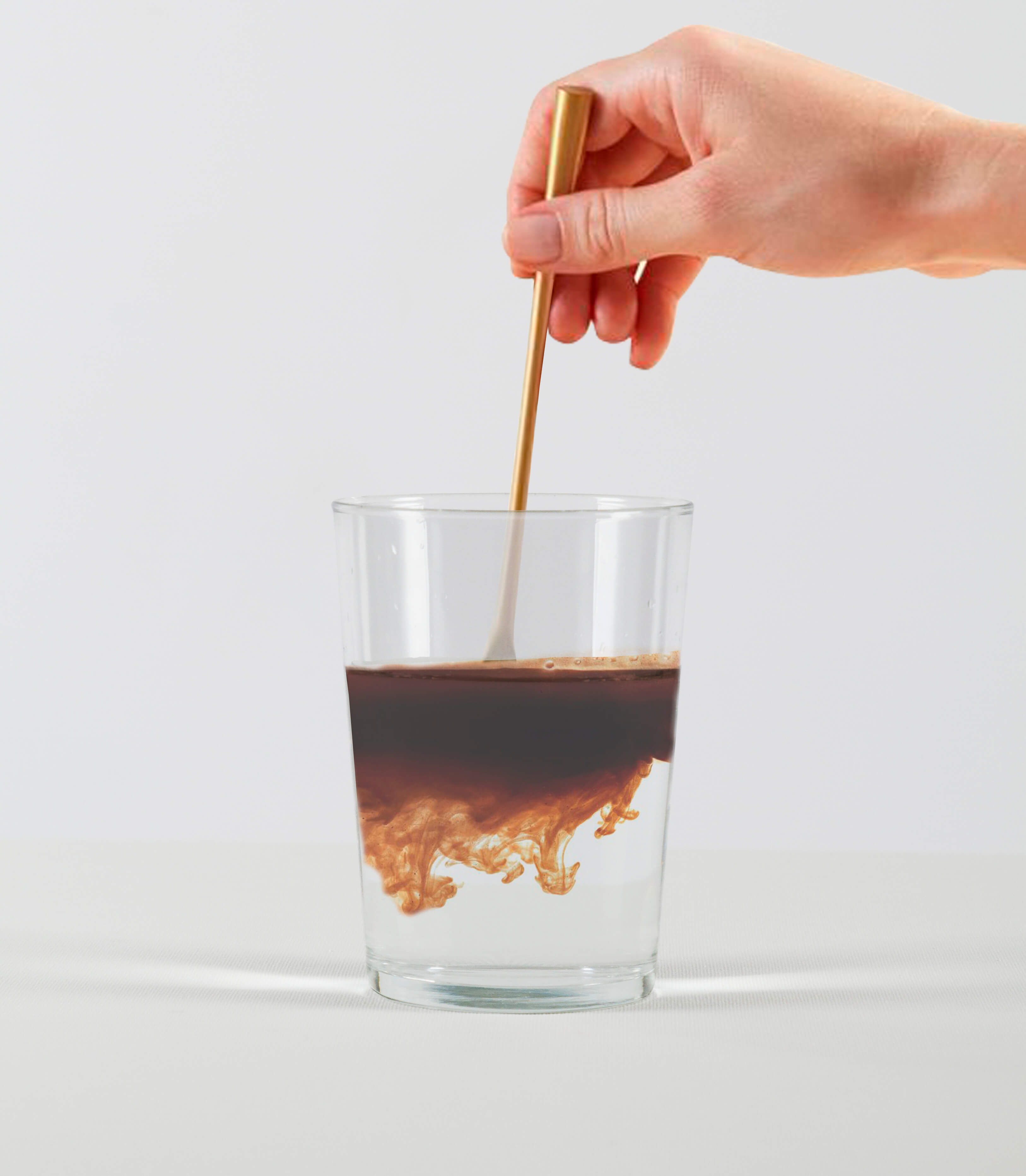 A hand stirs a dark liquid into a glass of water with a long, thin gold spoon.