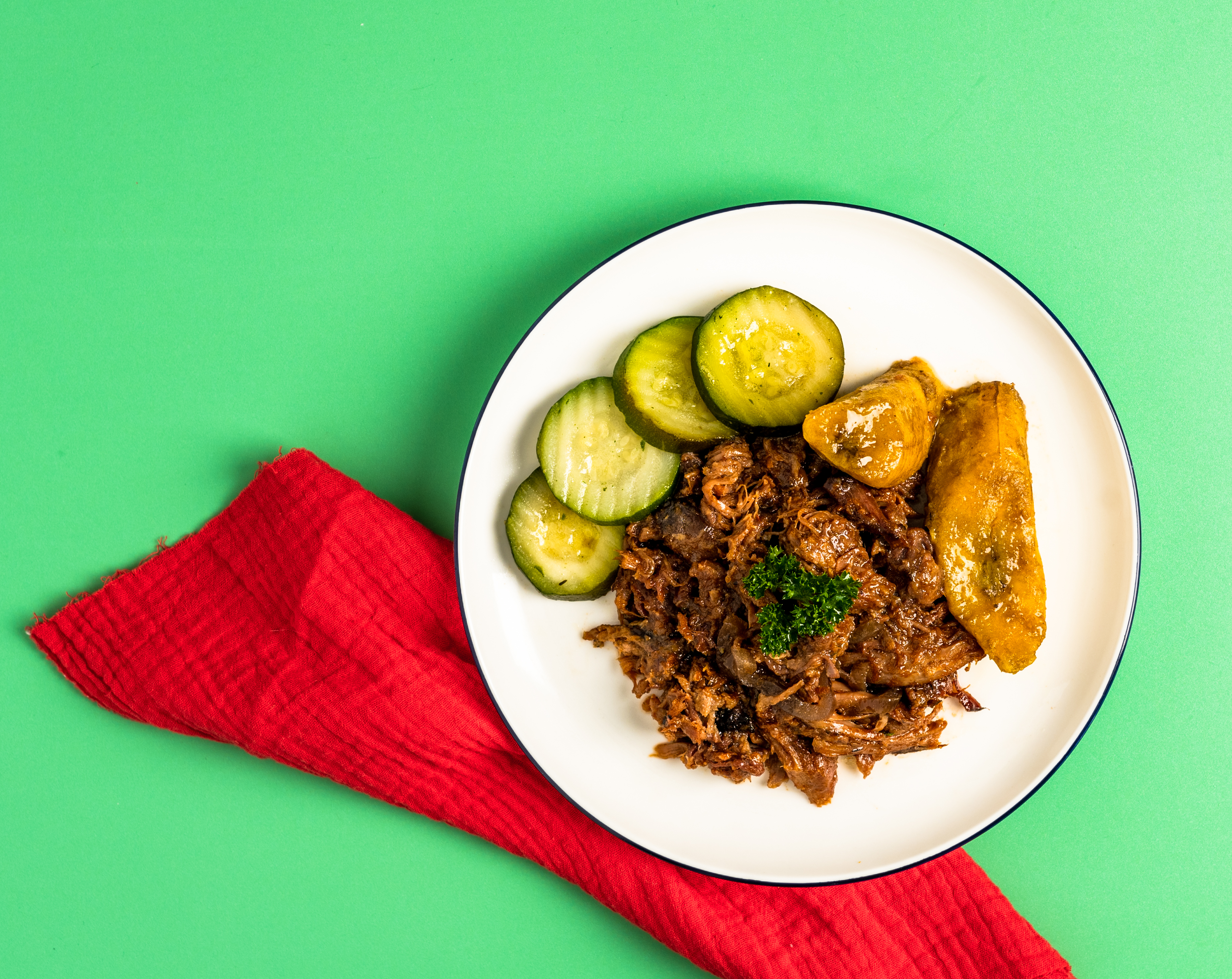 Plate of food with meat, cucumbers, and plantains on a green background with red cloth.