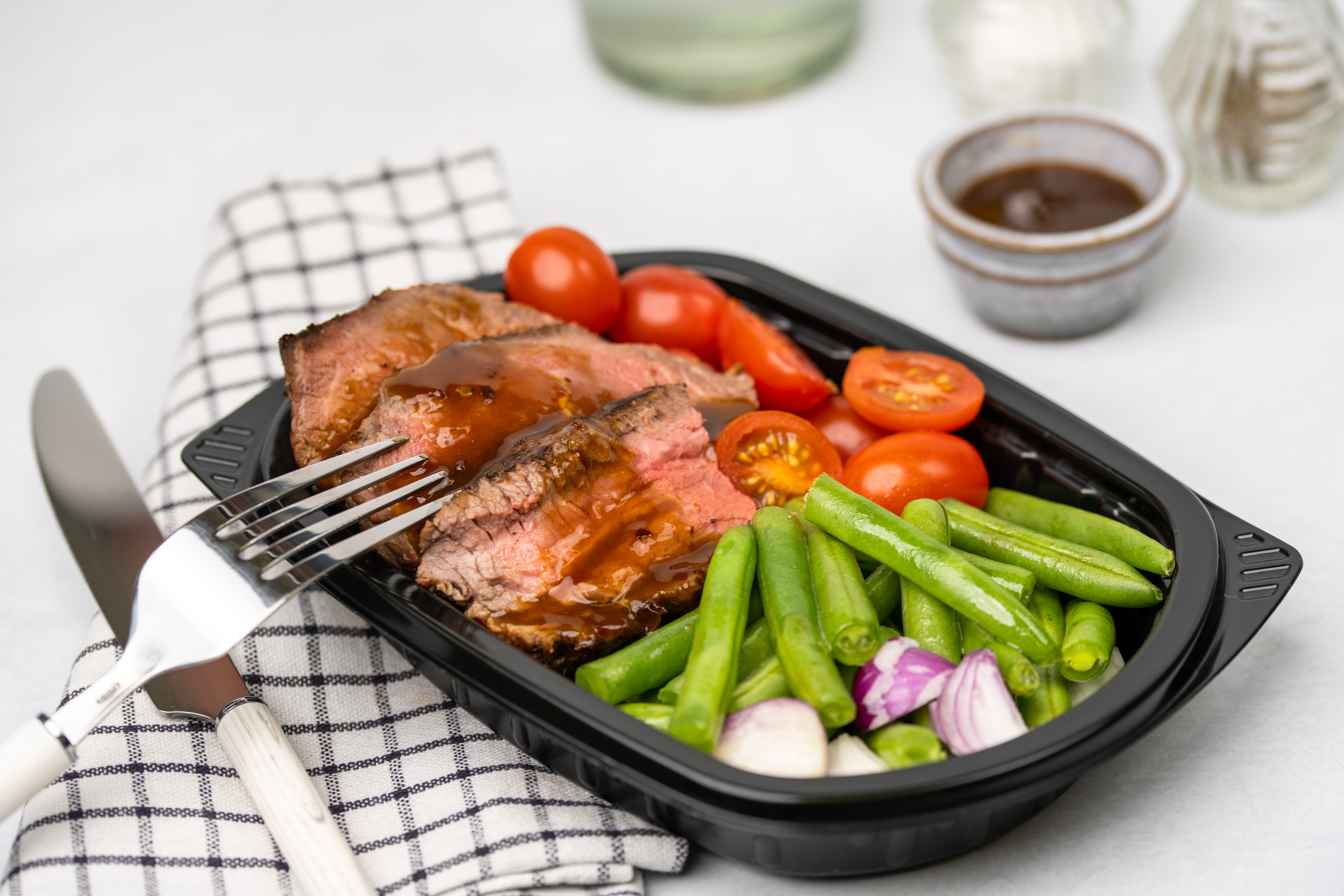 Sliced beef with sauce, cherry tomatoes, and green beans in a black tray.