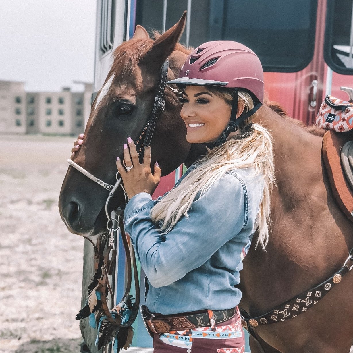Smiling person wearing helmet embraces a brown horse beside a building.
