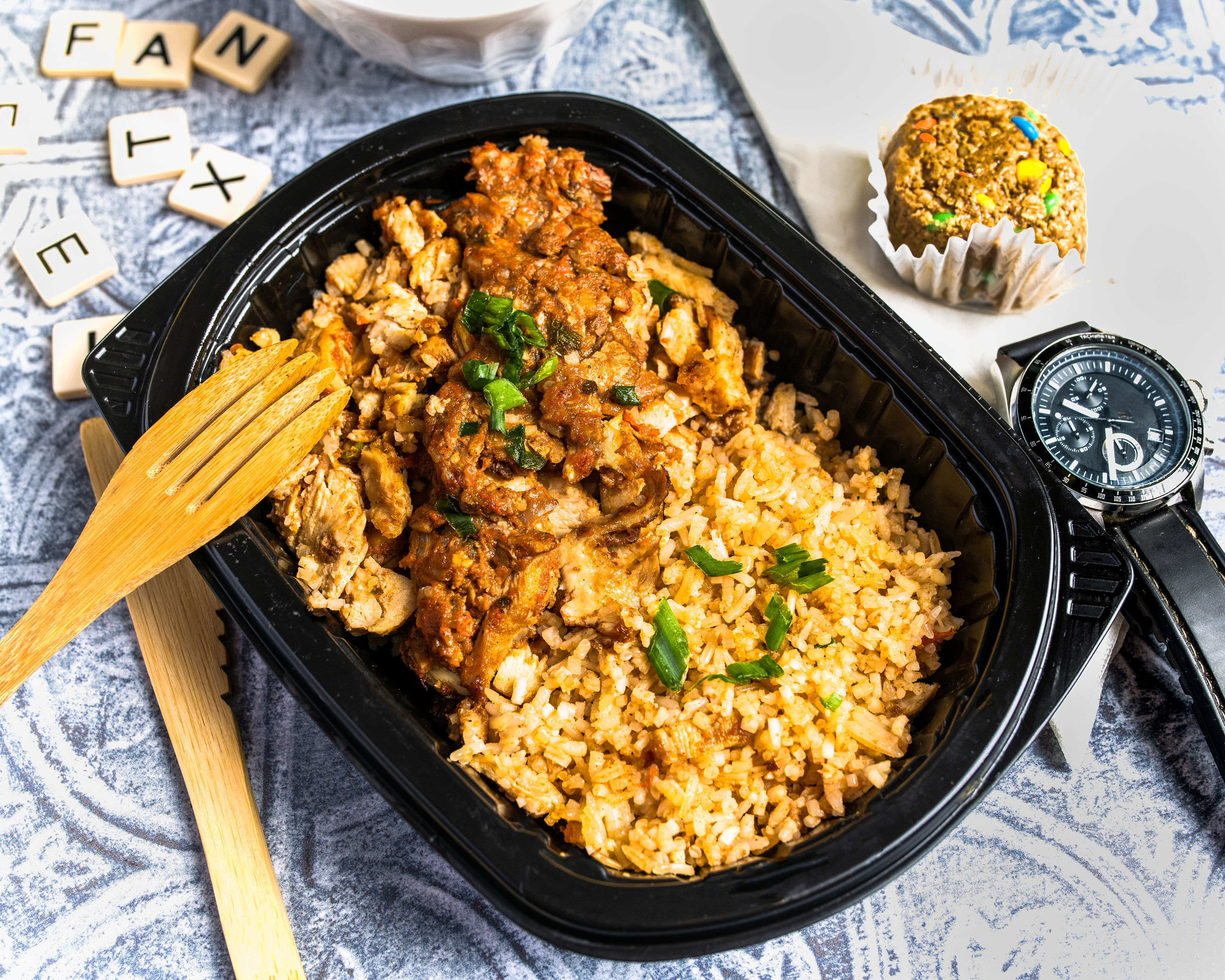 Black container with rice and chicken, wooden fork, watch, and a small dessert.