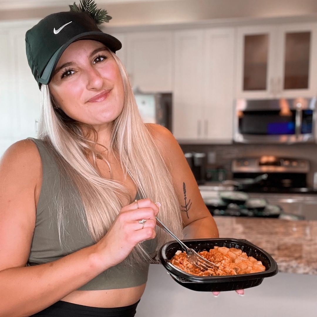 Person in a kitchen holding a bowl of food, wearing a cap and tank top.