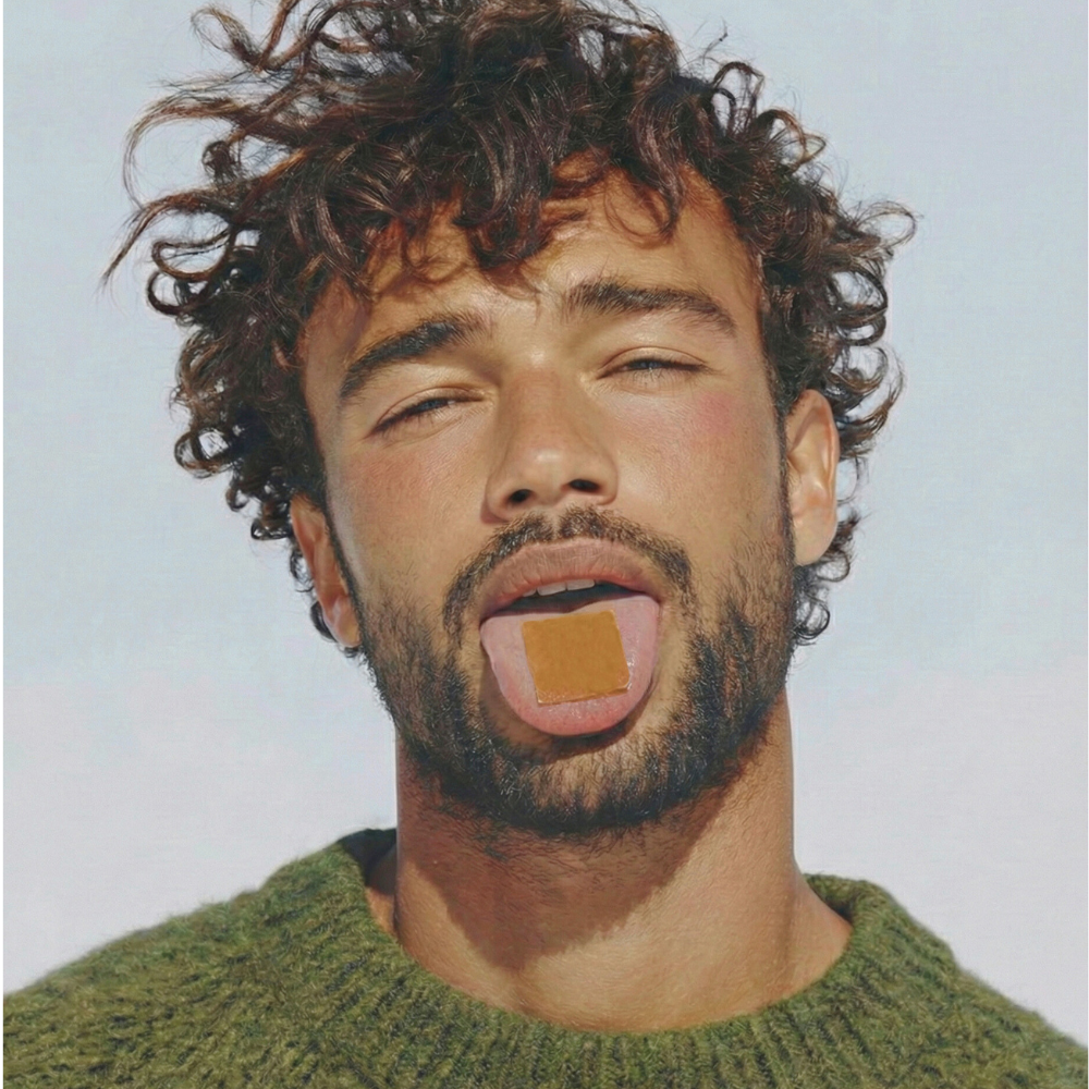 A close-up of a man with curly hair sticking his tongue out with a small tan square on it.