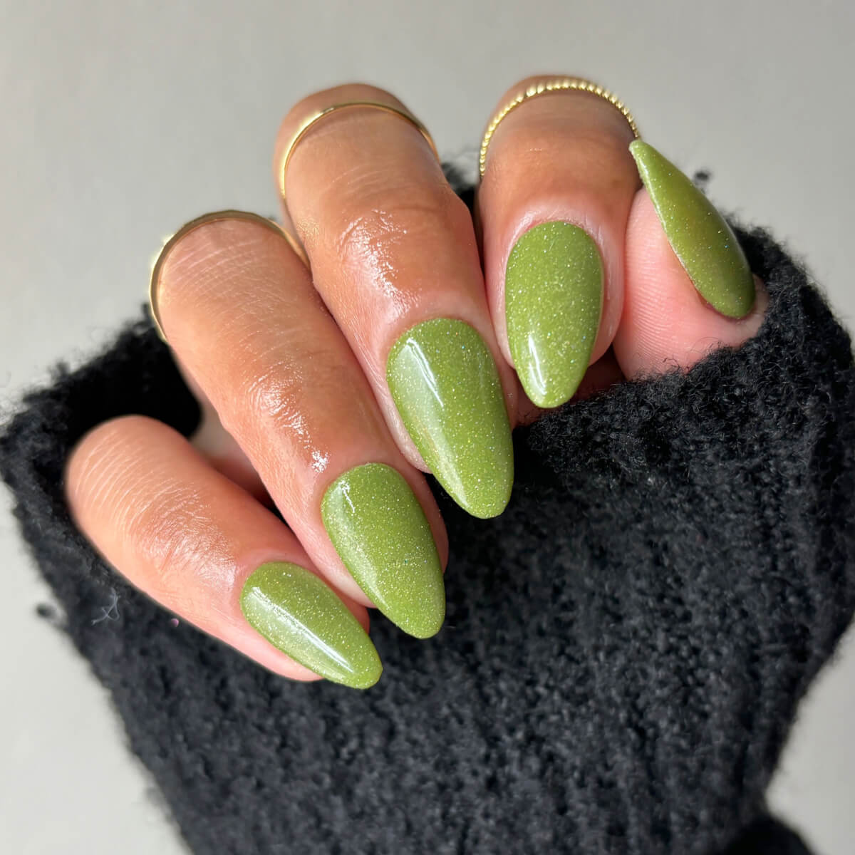 Hand with green glittery nails and gold rings, wearing a black sweater.