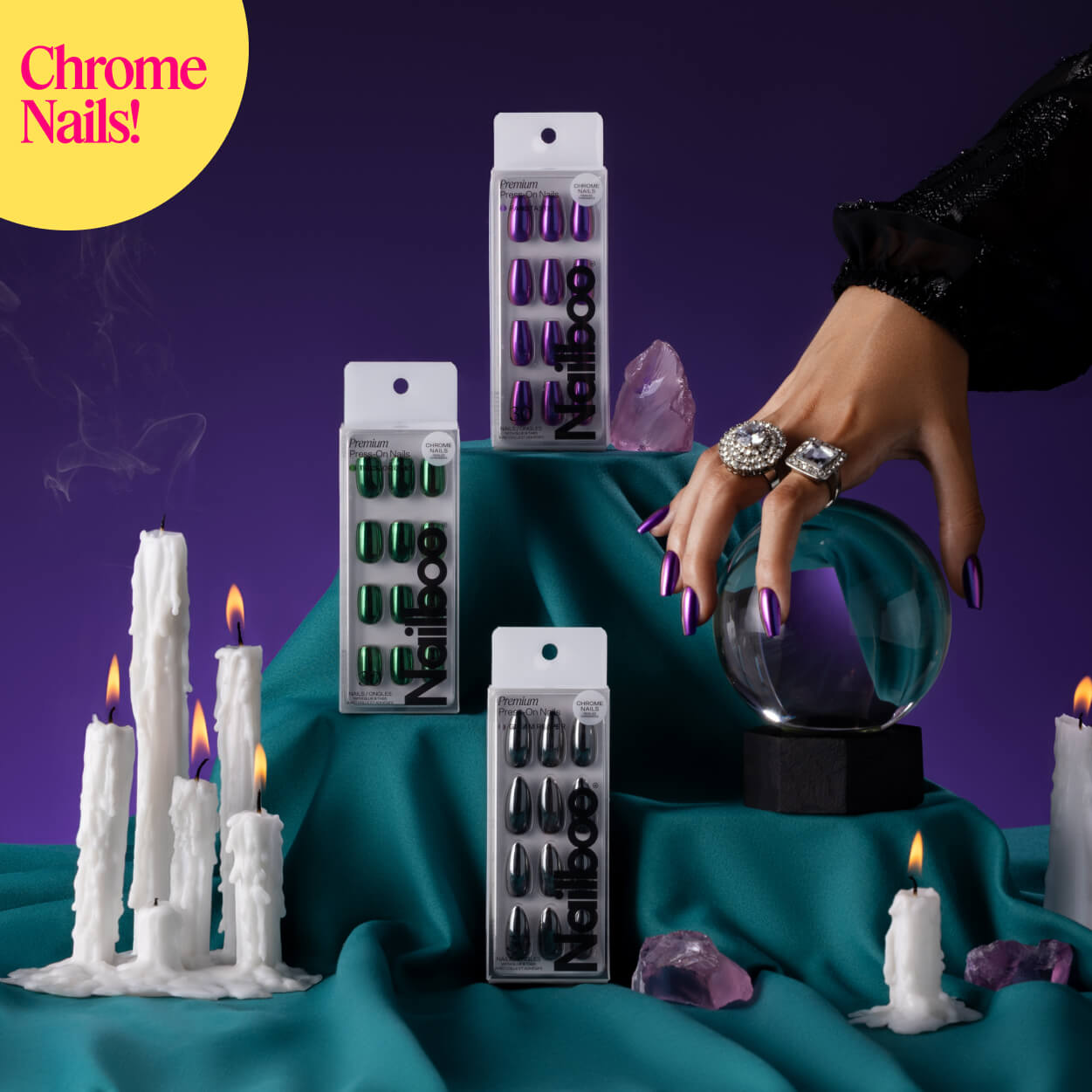 Hand with chrome nails on crystal ball, surrounded by candles and nail polish packages.