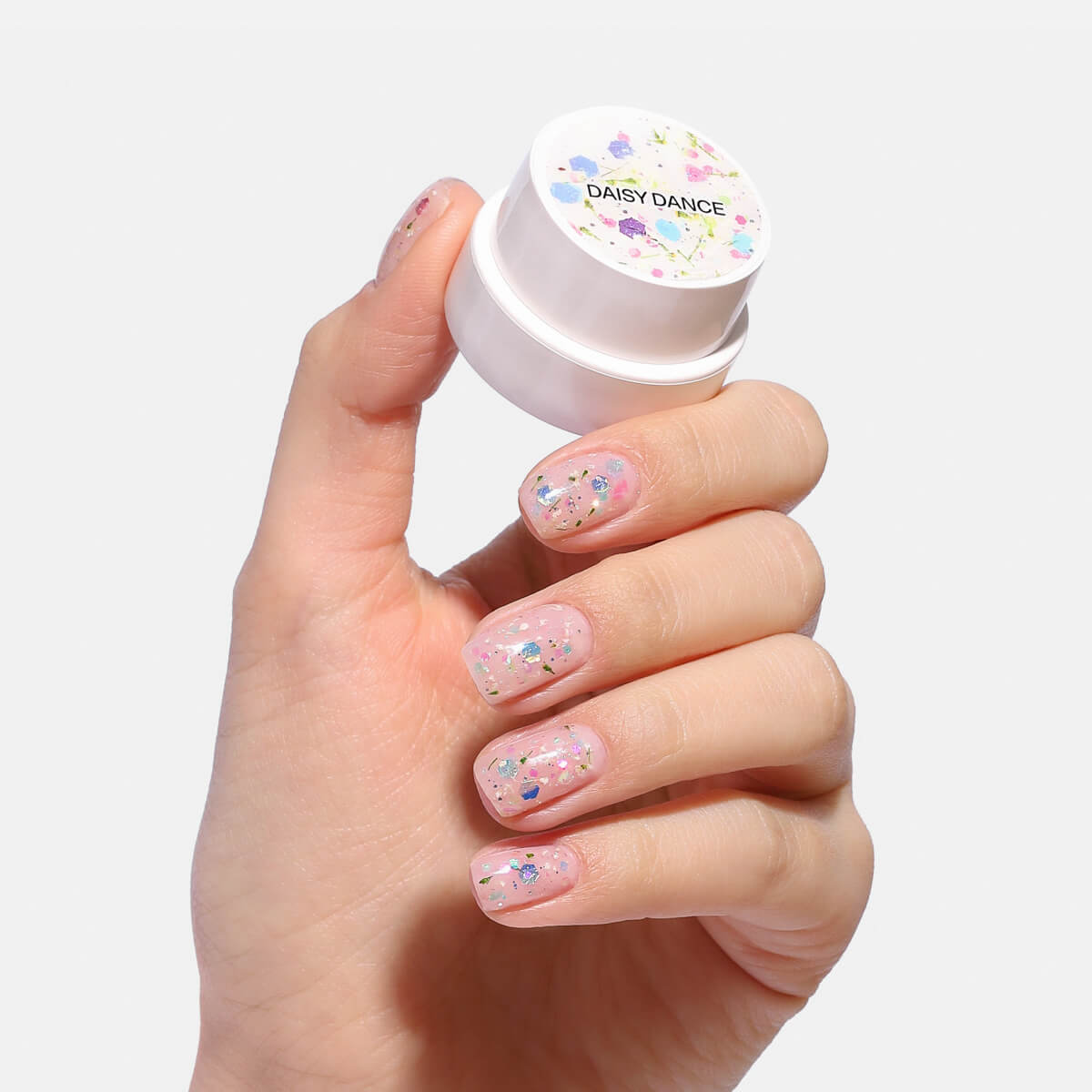 Hand with glittery manicure holding a 'Daisy Dance' nail product container.