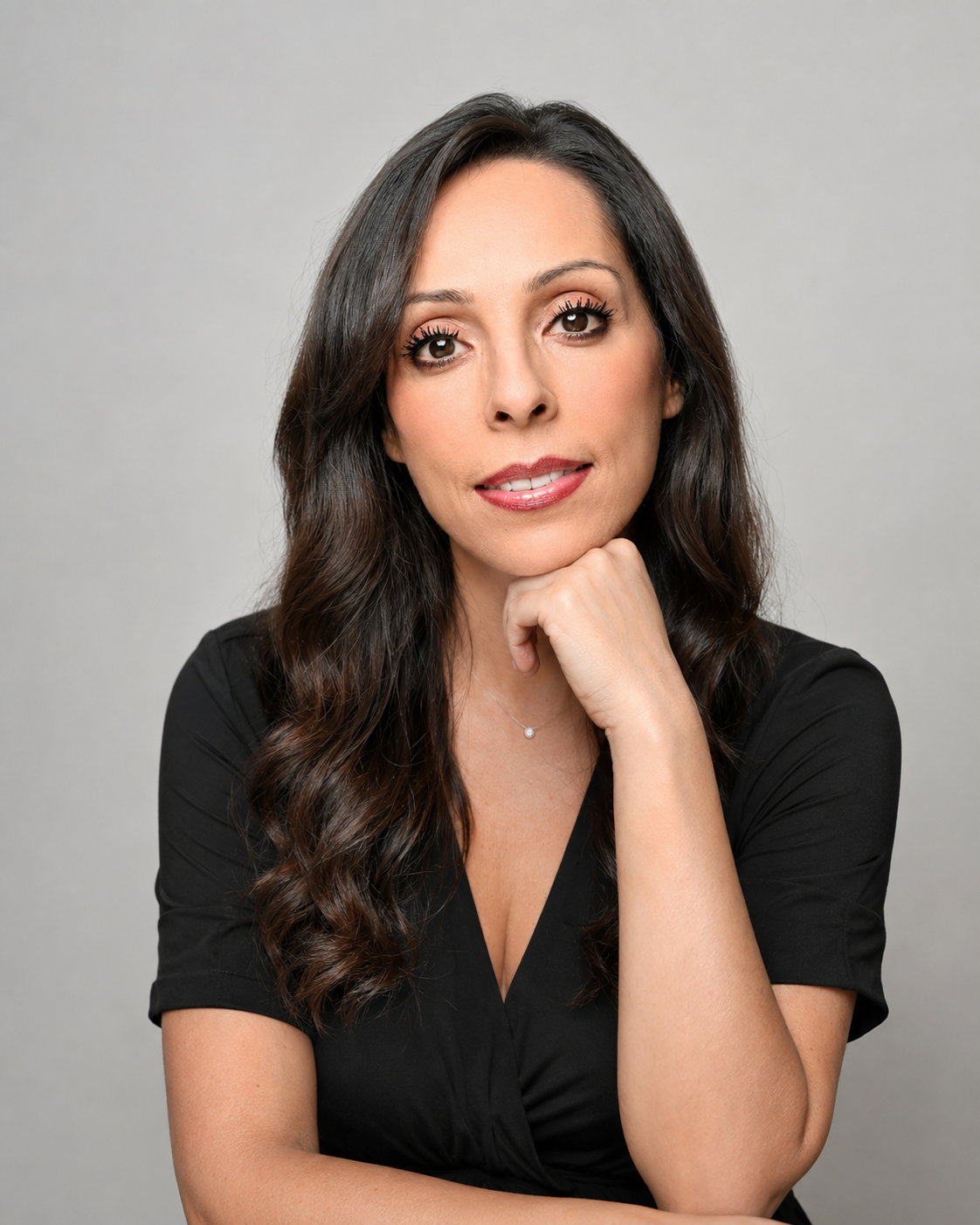 A professional headshot of a woman with long dark hair resting her chin on her hand against a gray background.