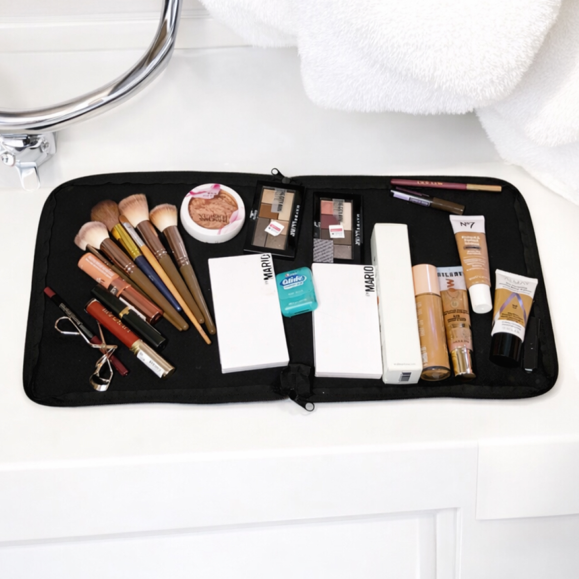 Various makeup products and brushes are laid out on a black mat on a white bathroom counter.