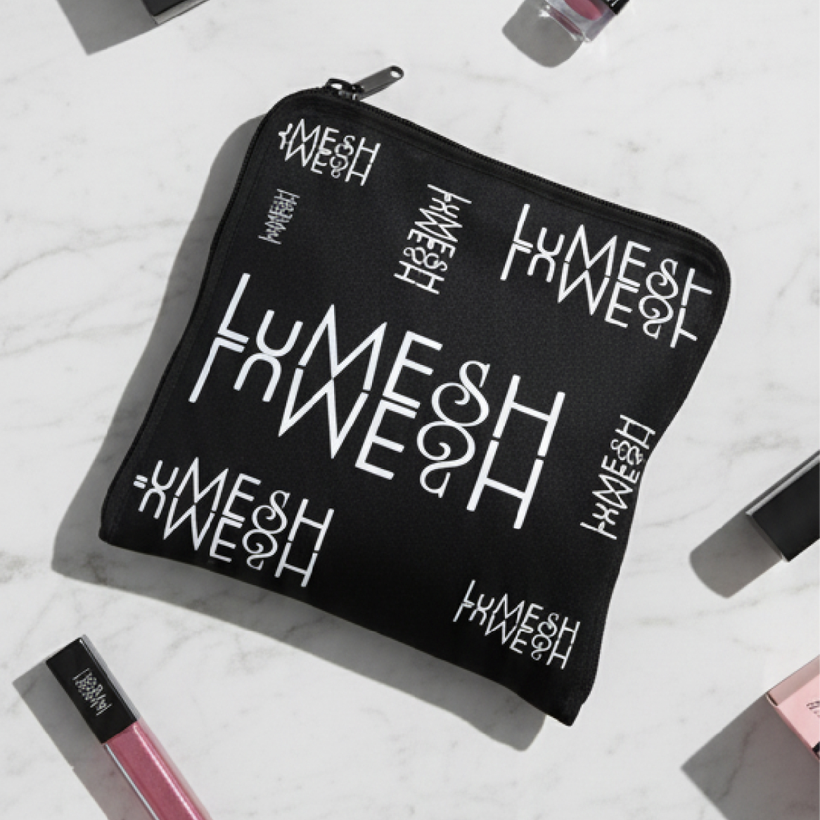 A black pouch with a repeating white logo pattern sits on a marble surface with makeup items.