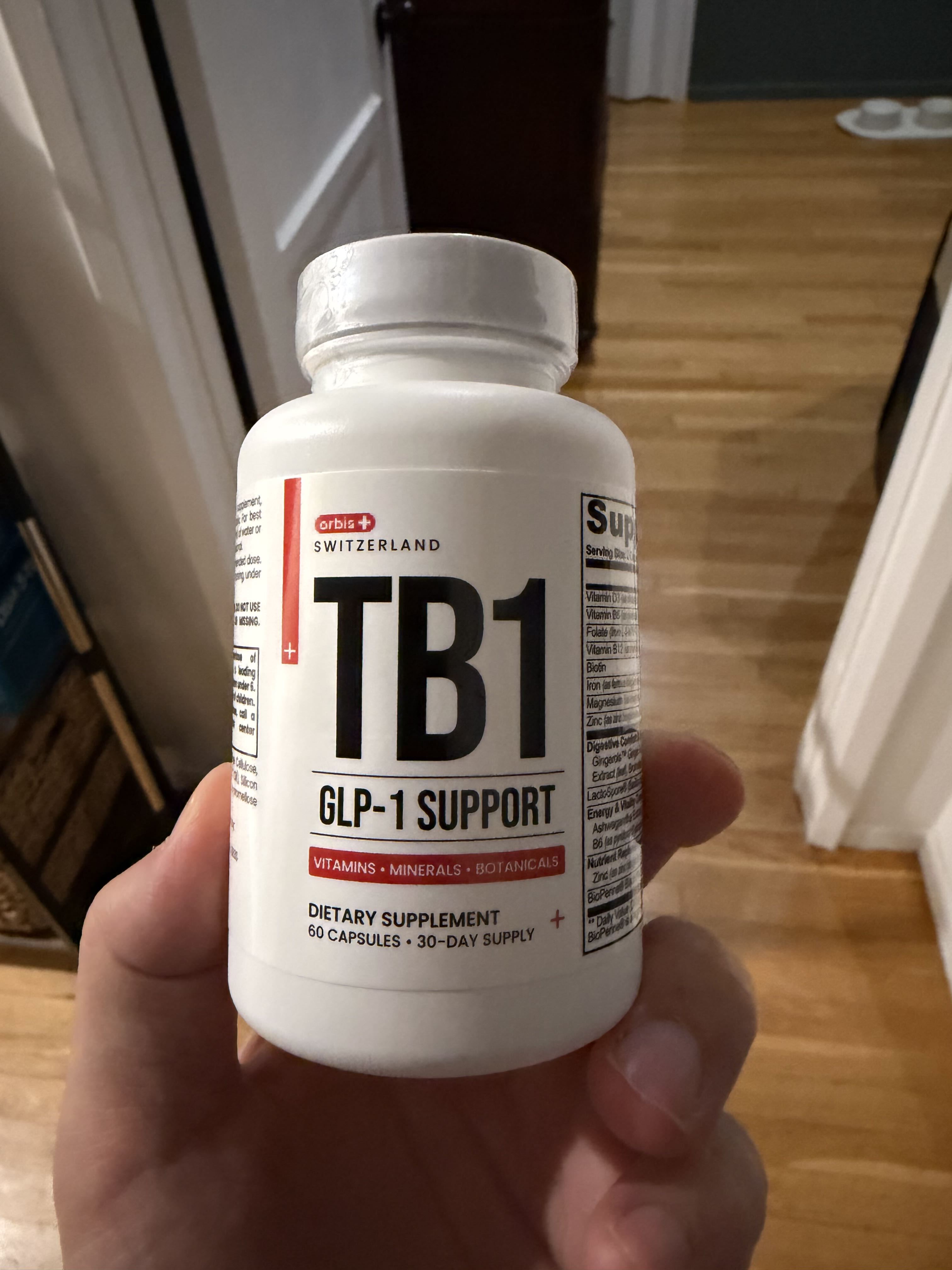A hand holding a white bottle of Orbis+ TB1 GLP-1 Support dietary supplement against a wooden floor.