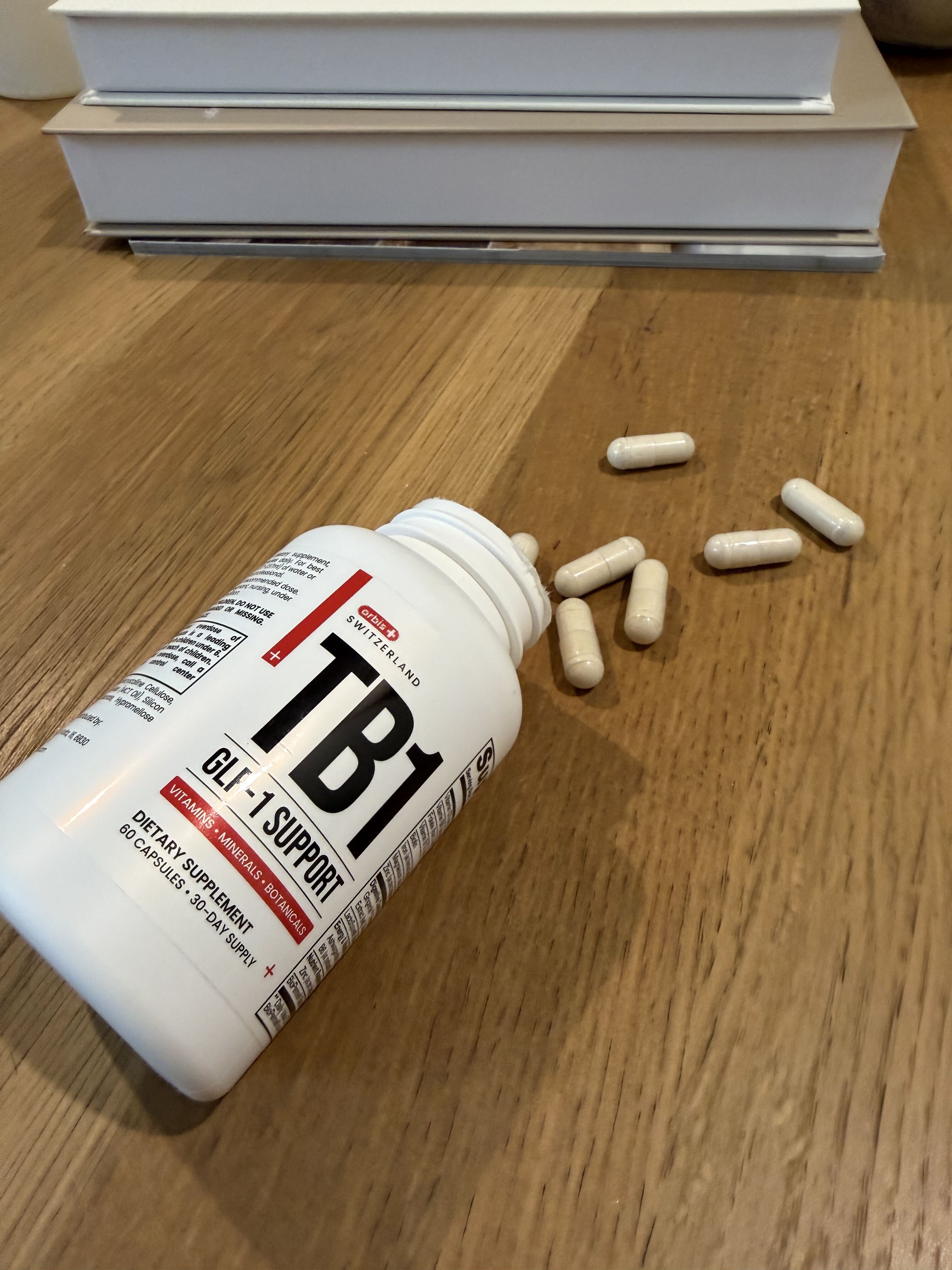 A bottle of TB1 dietary supplements lies on its side with several capsules spilled out onto a wooden table.