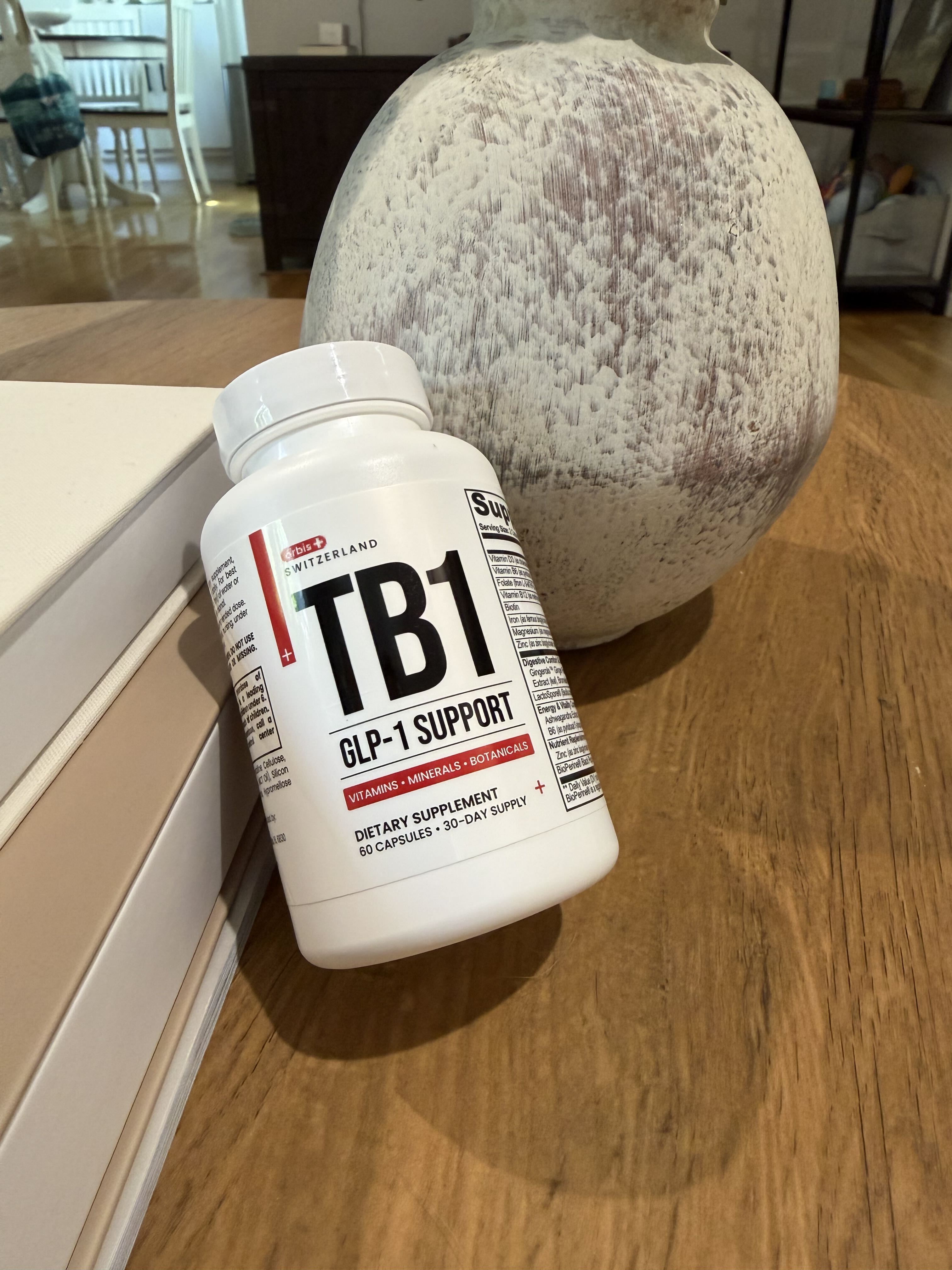 A white bottle of arbis+ TB1 GLP-1 Support supplements sits on a wooden table next to a vase.