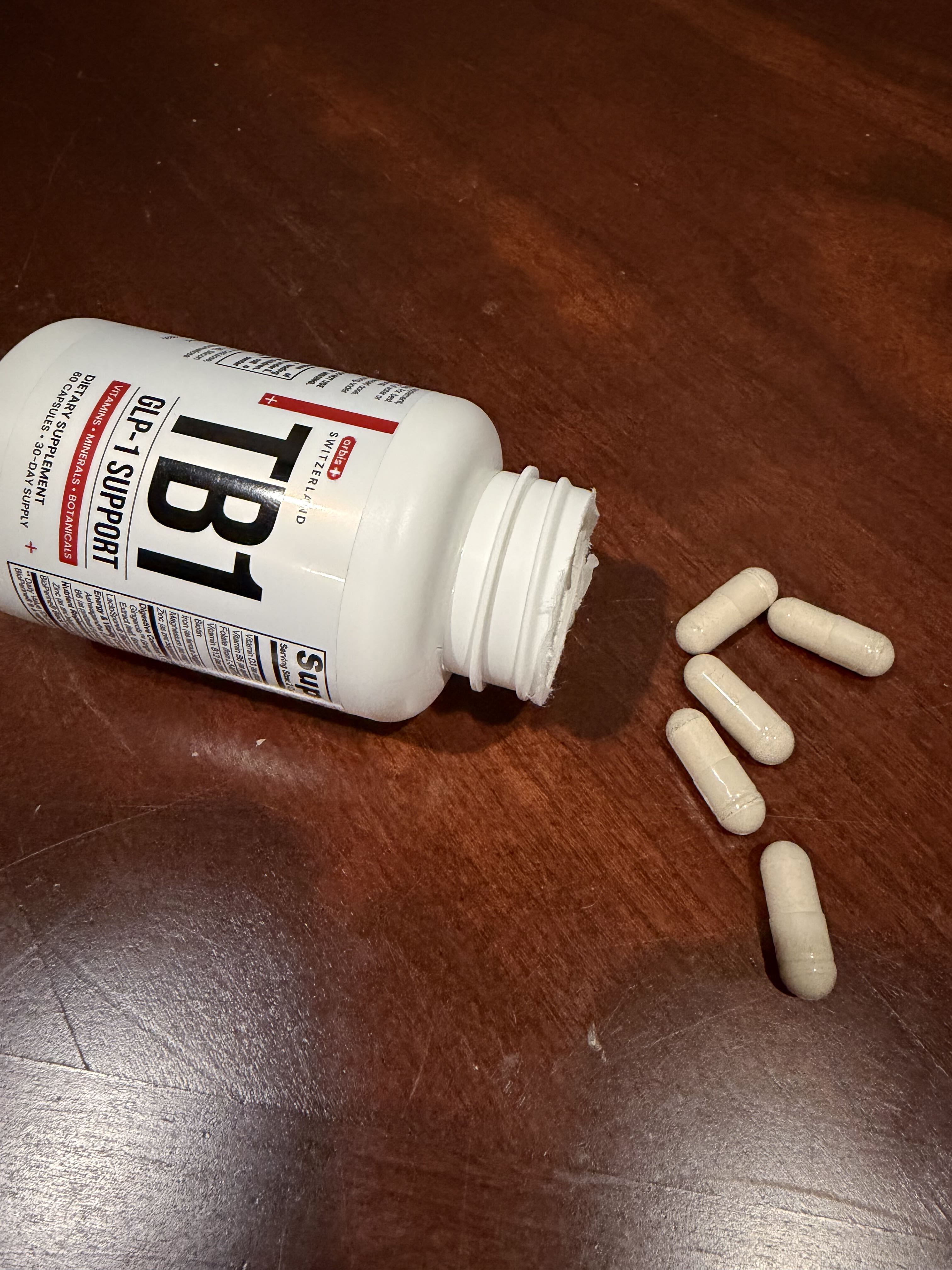 A white bottle of TB1 GLP-1 Support dietary supplement spilled on a wooden table with several capsules.