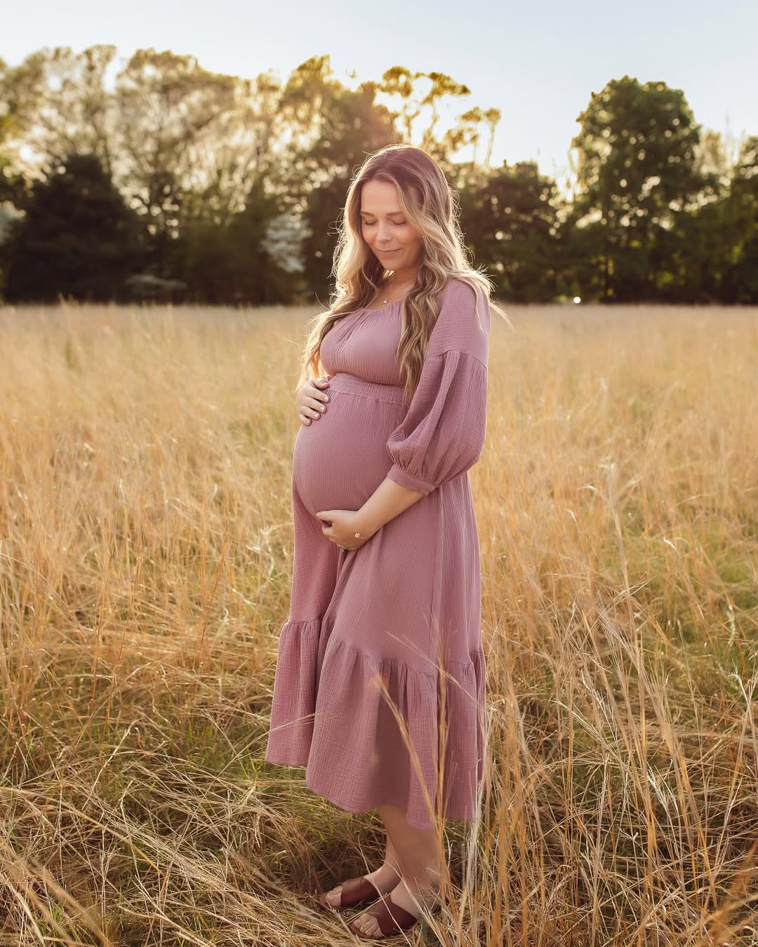 Pregnant woman in a field wearing a pink dress, holding her belly.