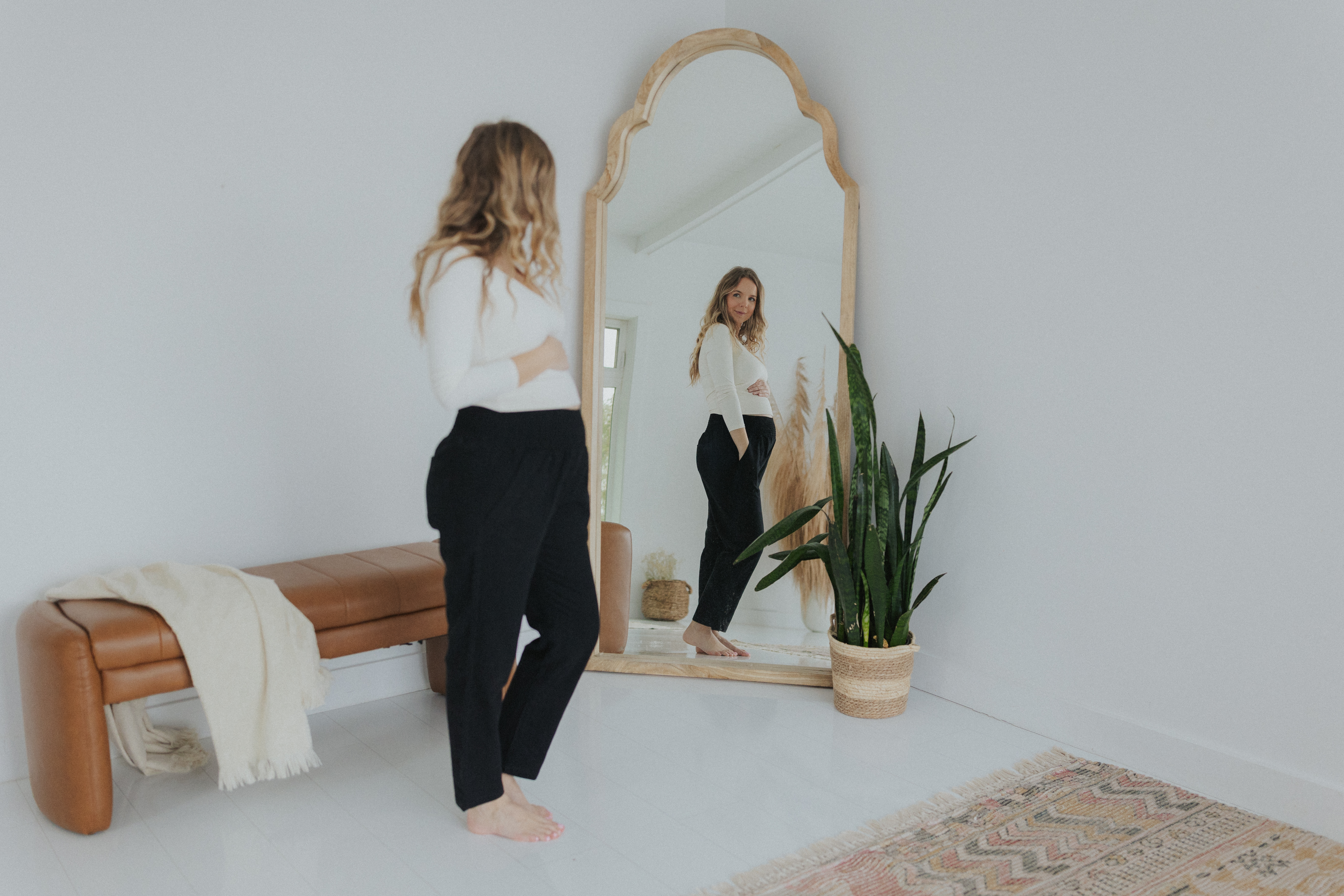 Woman in white top and black pants looking at mirror in a minimalist room.