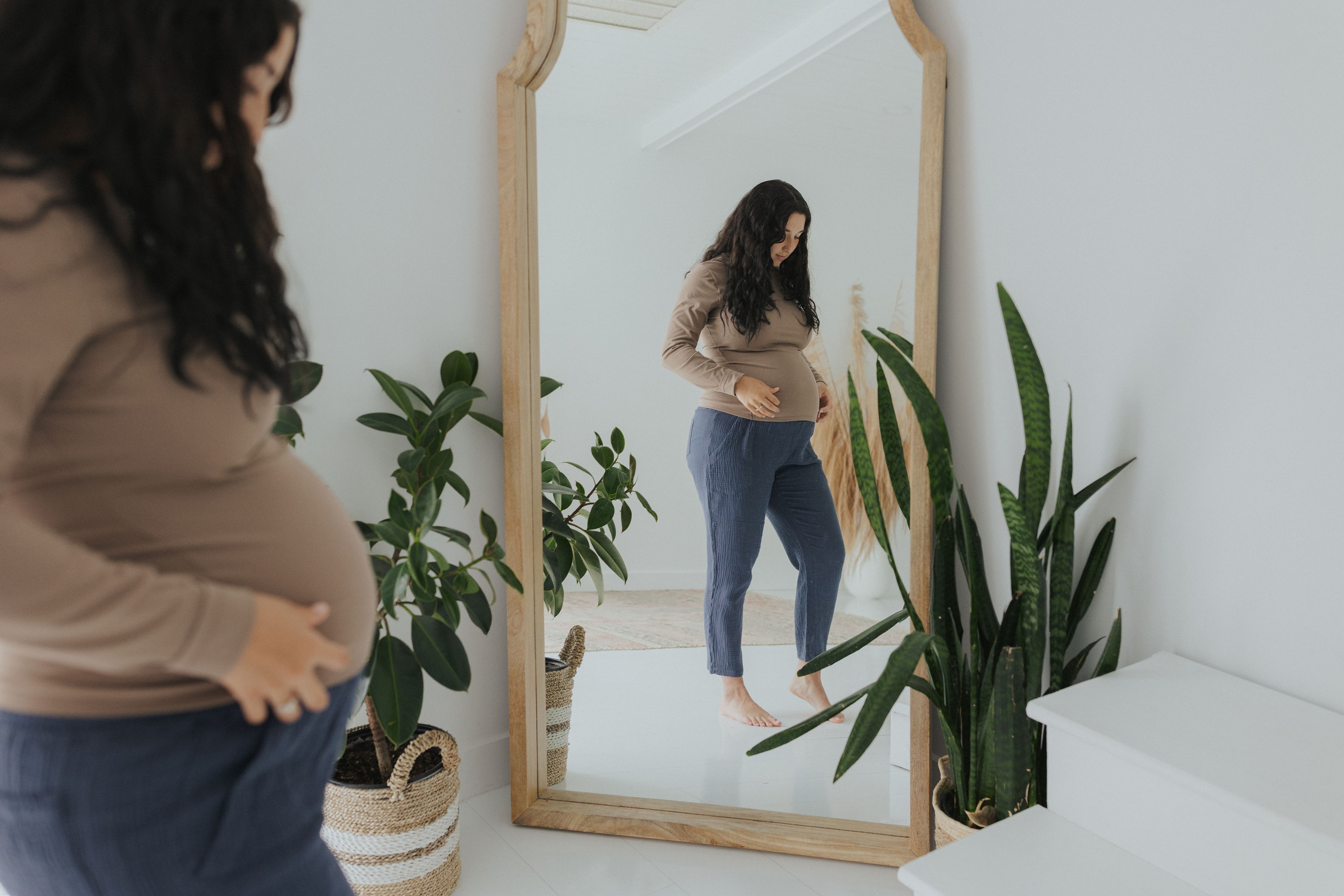 Pregnant woman looking at her reflection in a mirror, surrounded by plants.