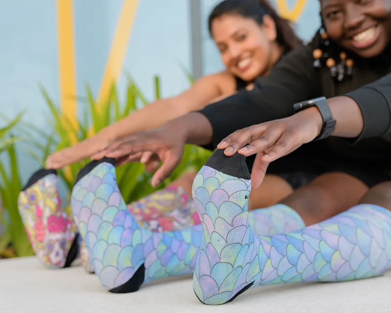Two smiling women stretch while seated, reaching for their toes in colorful socks with a scale pattern.
