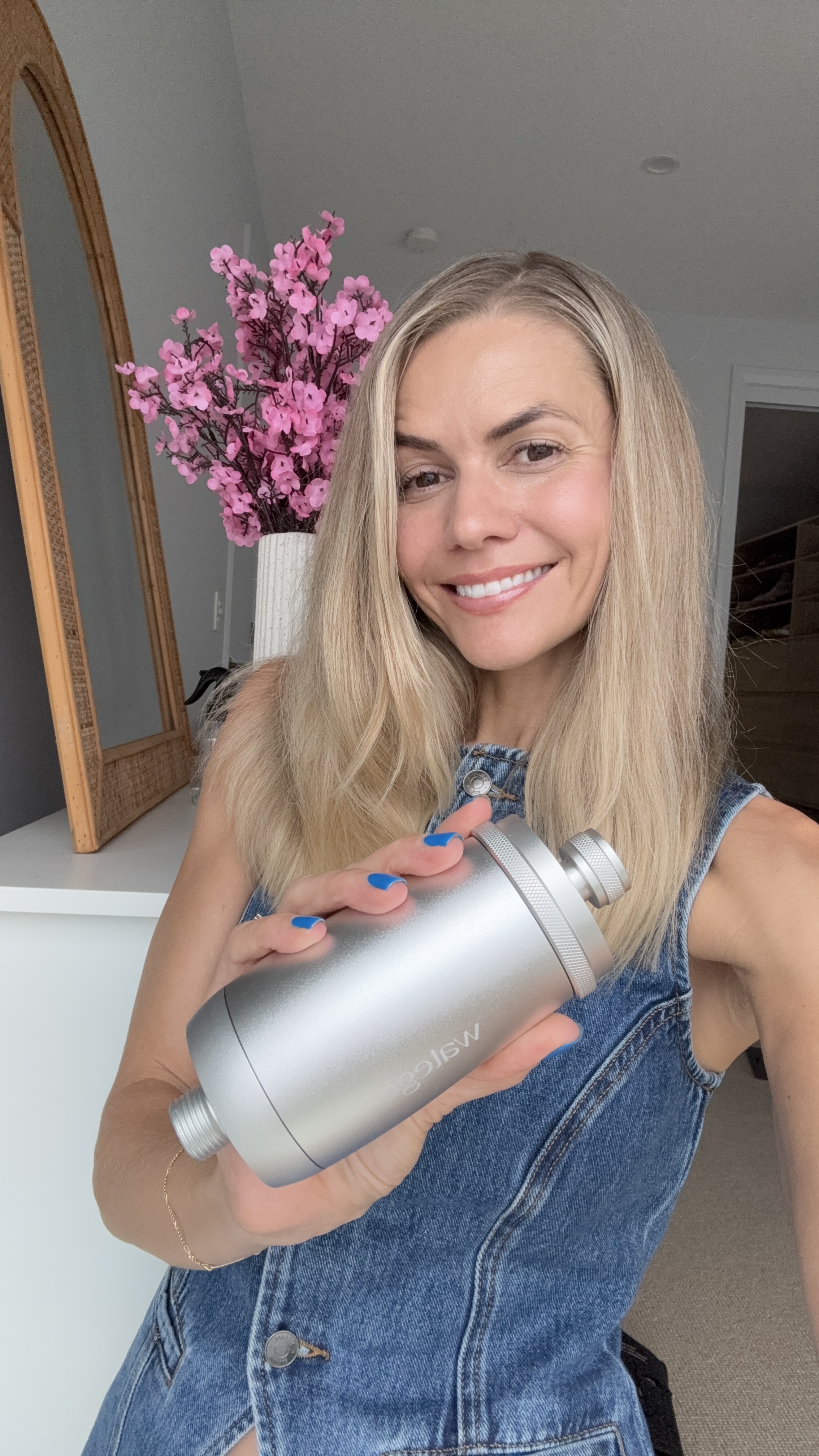 Person smiling and holding a metallic cylindrical object, with pink flowers in the background.