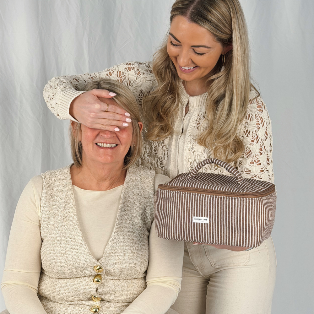 A woman covers a seated woman's eyes from behind while holding a striped bag, both smiling.