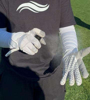 A person wearing white athletic gloves sprays a mist from a bottle onto the palm of their hand.