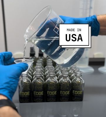 A person in blue gloves pours liquid from a beaker into rows of small glass bottles labeled 'EDGE'.