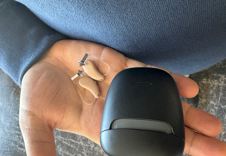 A person's hand holding a pair of hearing aids and a black charging case.