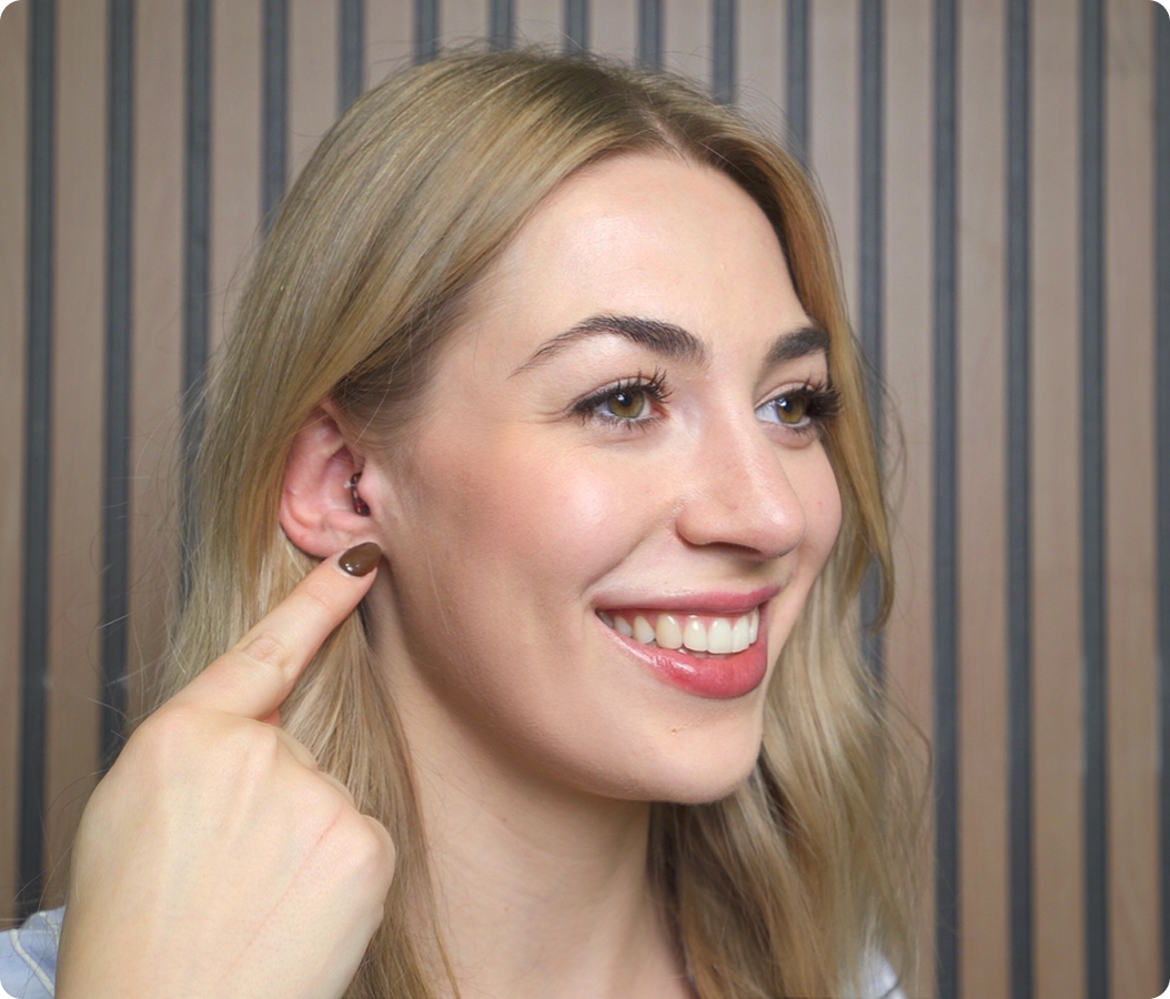 Smiling person pointing to ear with ear piercing, wood panel background.