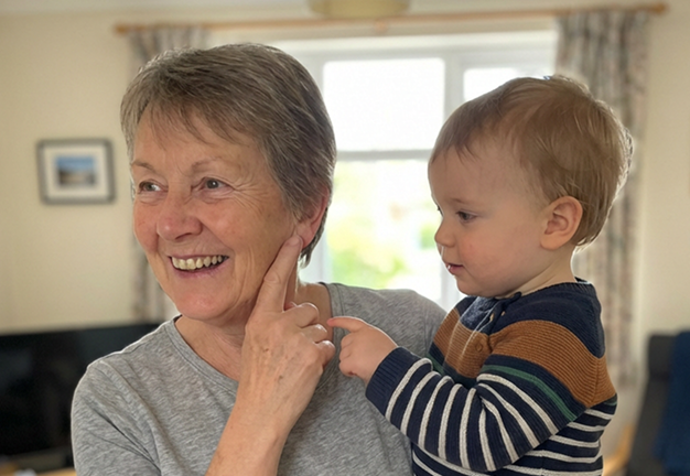 An older woman with short gray hair smiles while a young child points at her ear.