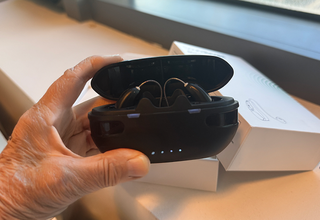 Hand holding a black wireless earbud case, open with earbuds visible.