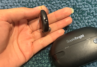 Hand holding a hearing aid next to a Soundbright case on a blue textured surface.