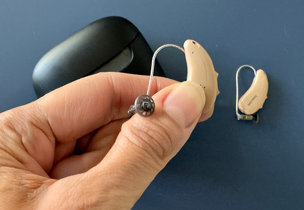 A hand holds a beige hearing aid, with another one and a black case on a blue surface.