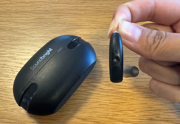 A hand holding an earbud next to its charging case on a wooden surface.