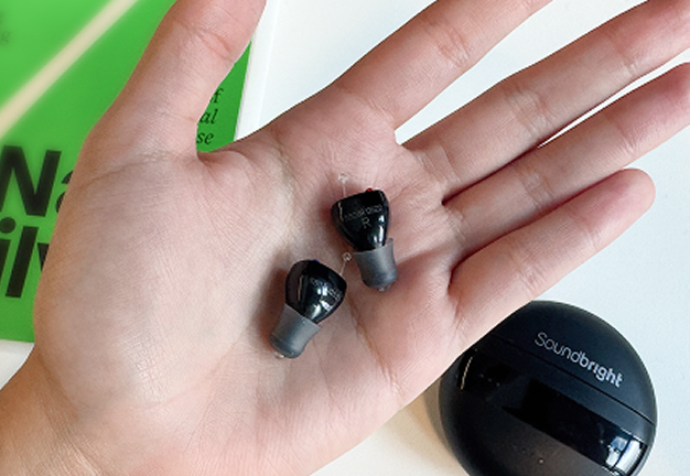 Hand holding two black earbud headphones, with a round black case nearby.