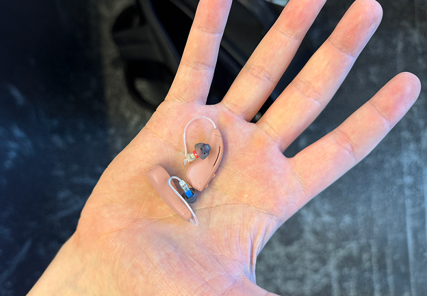 A hand holding two small hearing aids on a dark background.