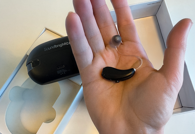 Person holding a black hearing aid with a charging case in the background.