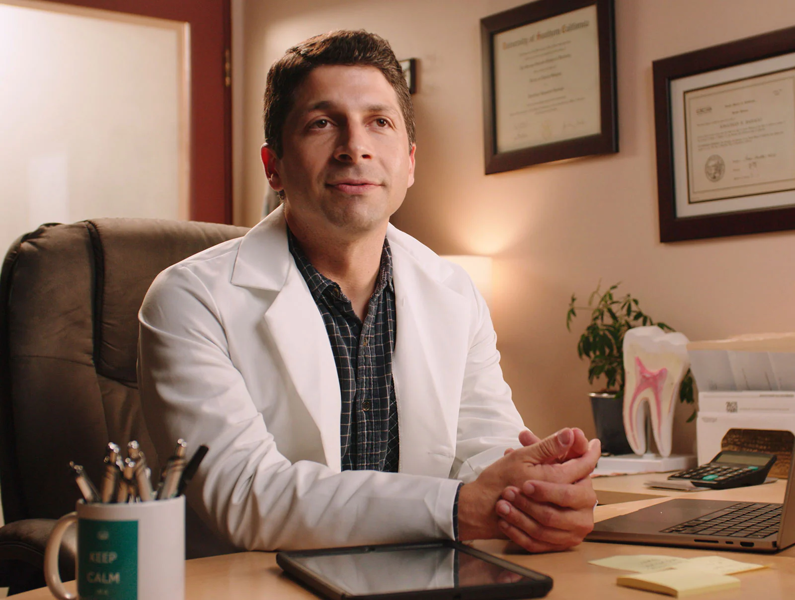 Man in white coat sitting at desk with documents and a plant in background.