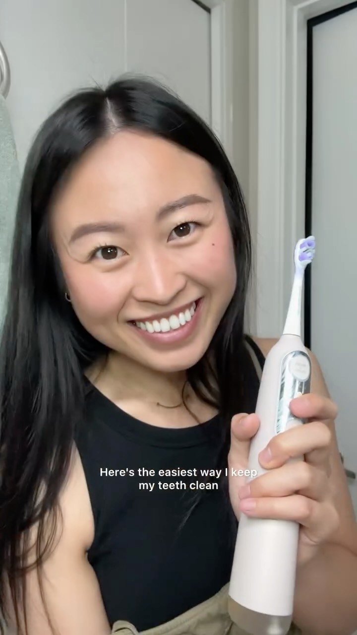 Person smiling and holding an electric toothbrush in a bathroom.