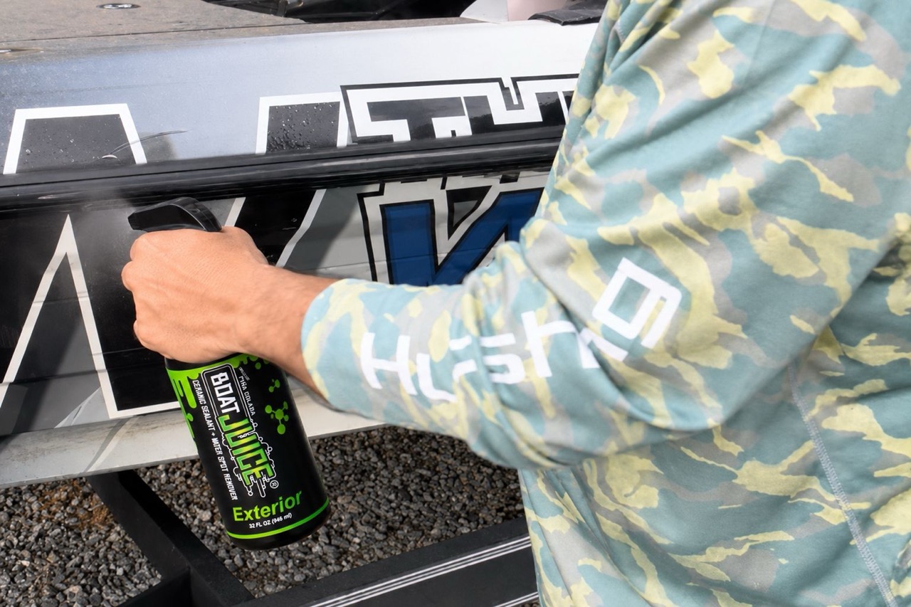 A person in a camouflage shirt sprays Boat Juice Exterior cleaner onto the side of a boat.