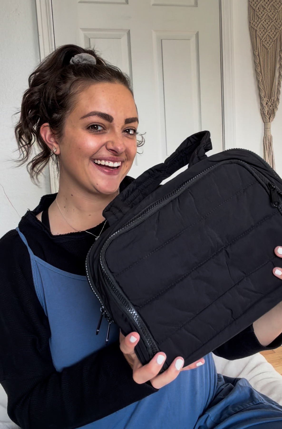 A smiling woman with her hair in a ponytail holds up a black quilted bag.