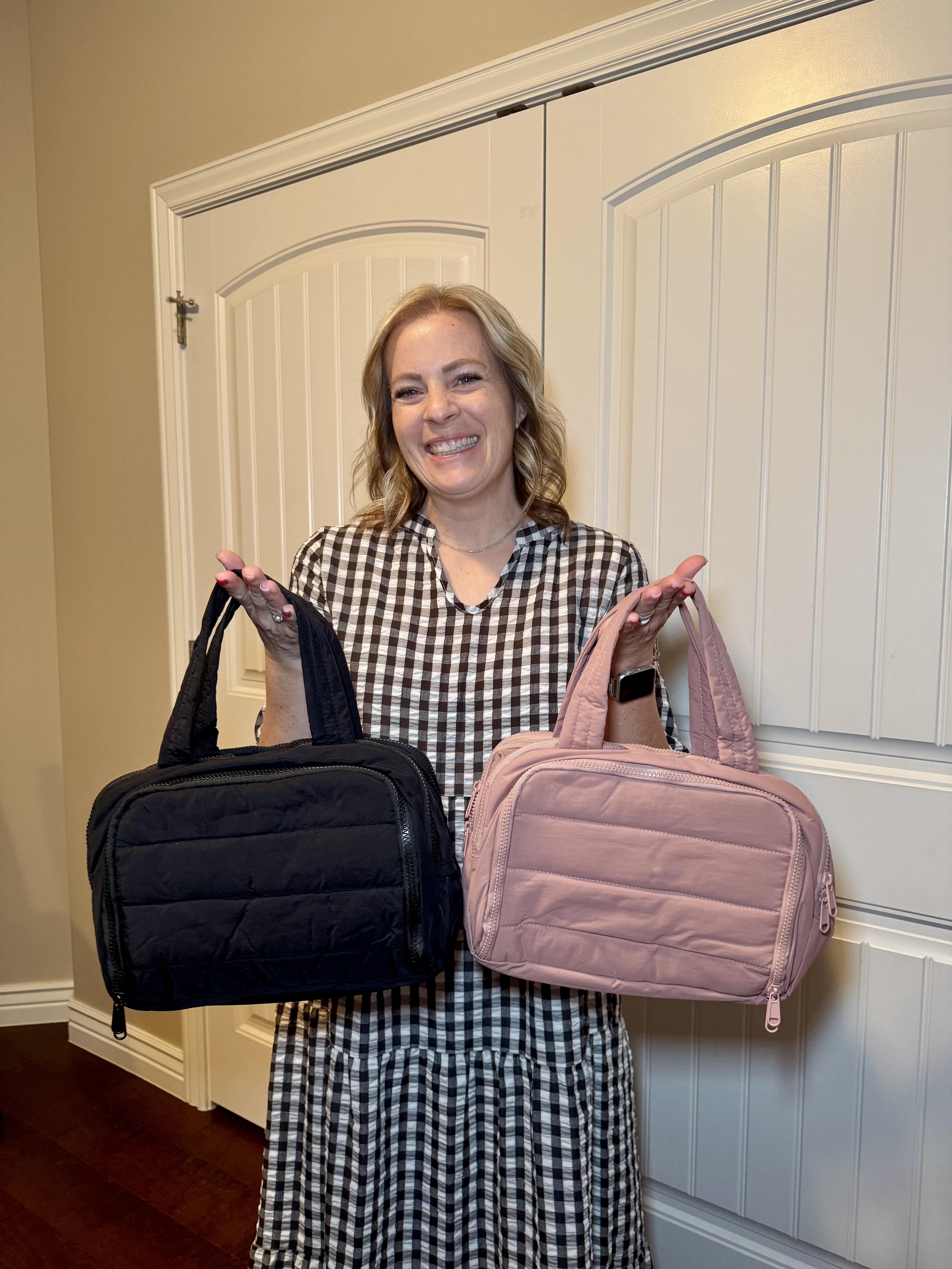 A woman in a checkered dress holds up two quilted bags, one black and one pink.