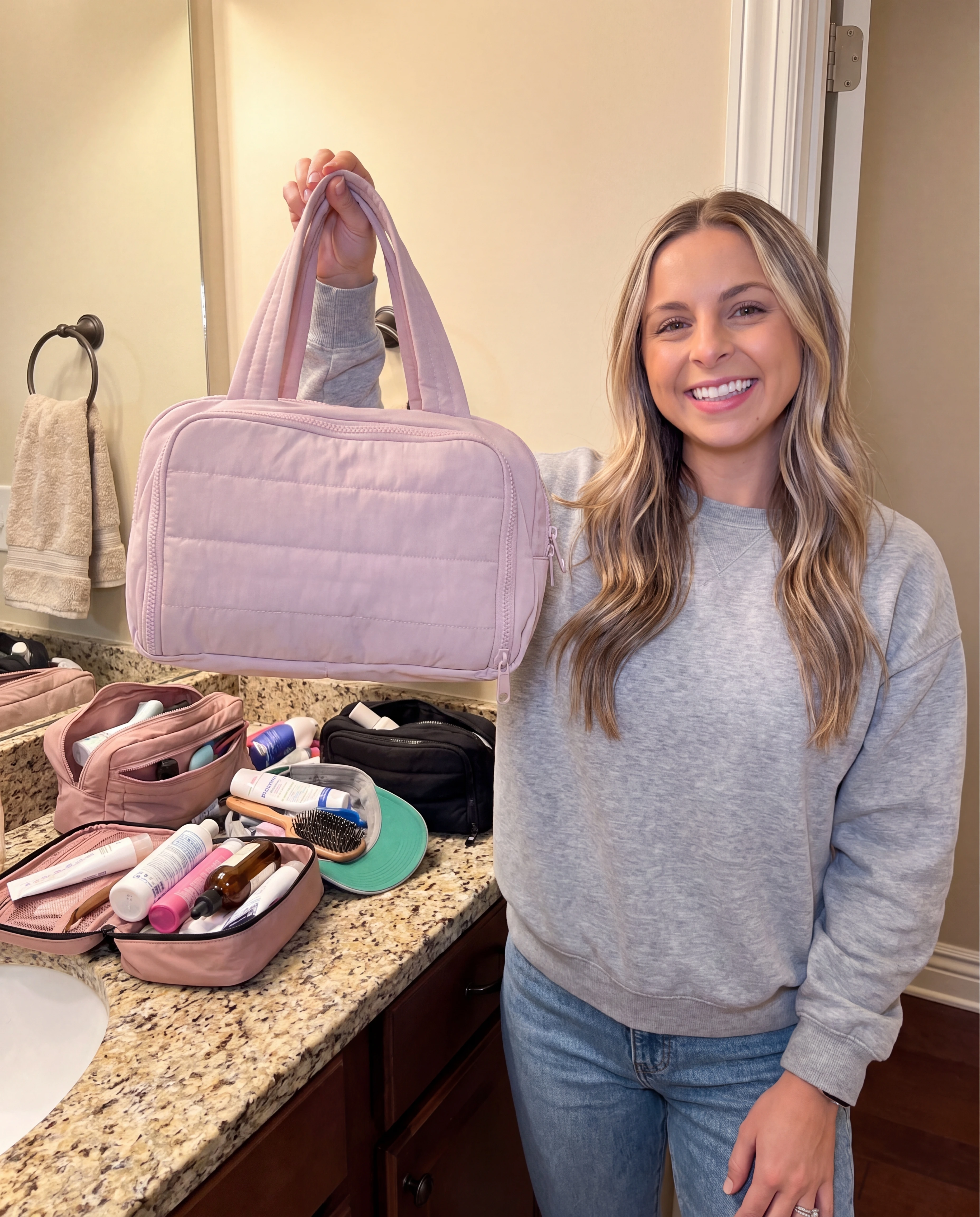 A woman holds up a pink quilted bag in a bathroom with toiletries on the counter.