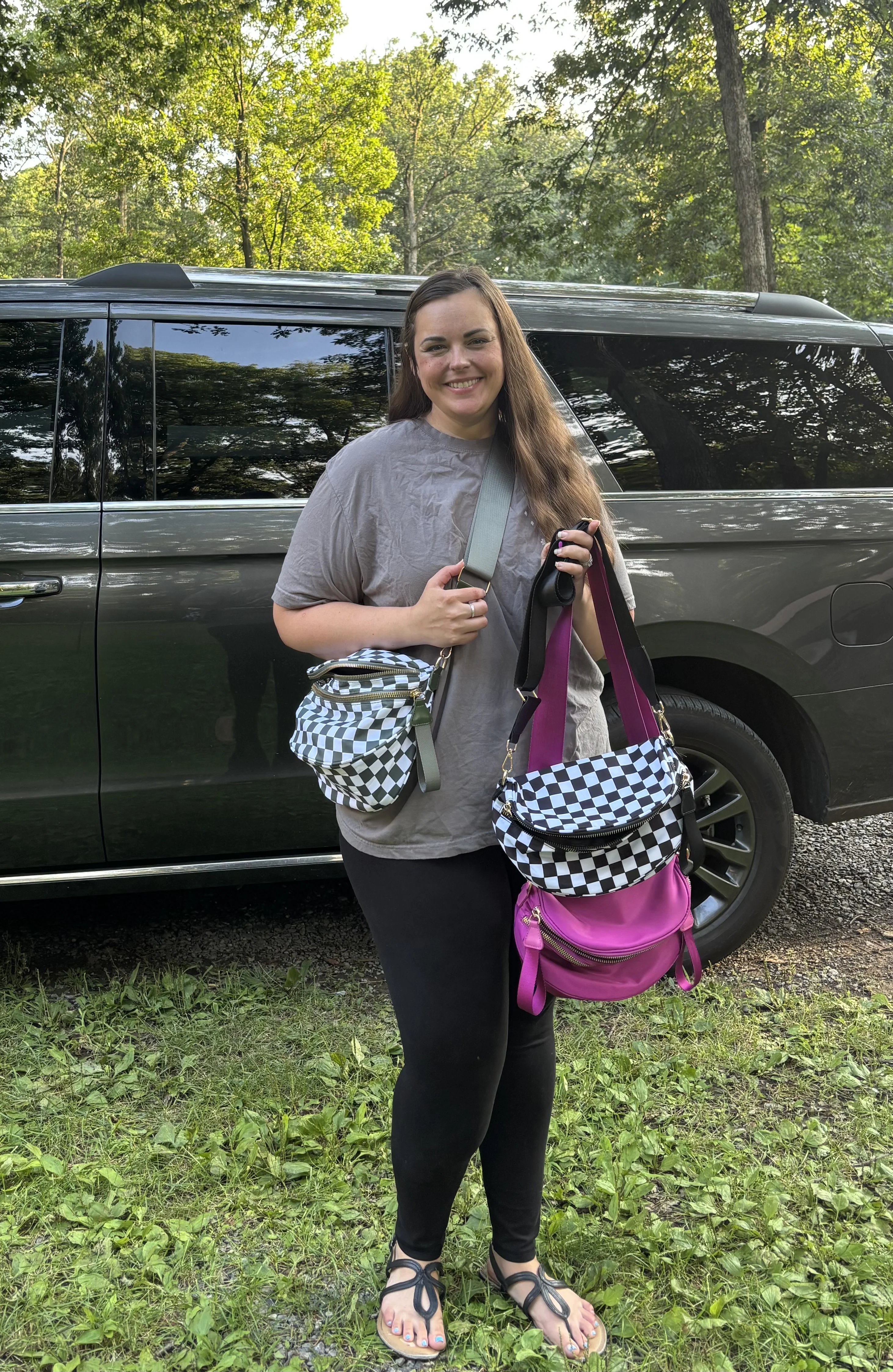 Person holding checkered and pink bags, standing by a car in a wooded area.